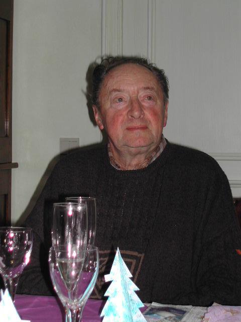 An older man sits at a table with empty glasses and small paper Christmas trees in front of him.