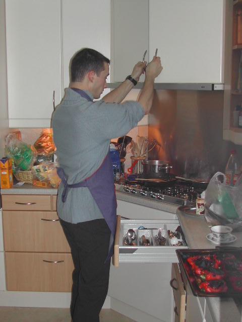 A man wearing an apron cooks in a kitchen, holding utensils while standing near a stove with food.