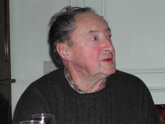An older man in a dark sweater sits at a table, looking to the side and speaking.