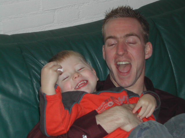 A man and a child laugh together on a green couch, with the child holding a small white object.