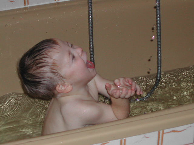 A young child in a bathtub reaches for water dripping from a shower hose, smiling with excitement.
