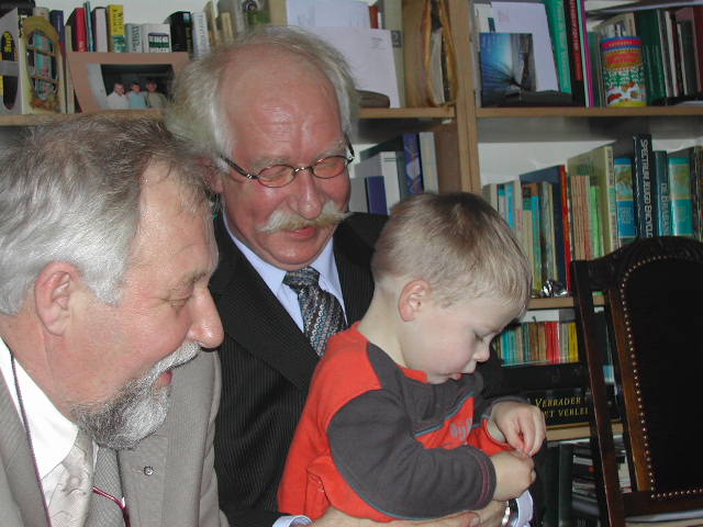 Two older men smile while a young child in a red sweater examines a small object in their hands.