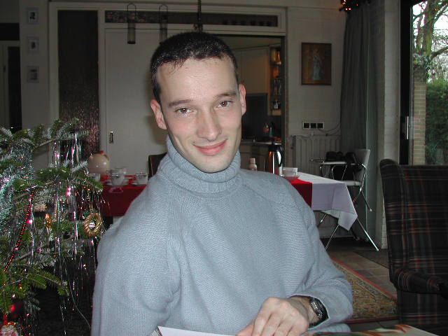 A man in a light blue sweater smiles while sitting near a decorated Christmas tree and a festive table.