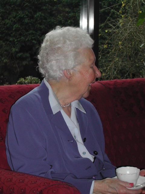 An elderly woman in a purple blazer sits on a red couch, holding a teacup and smiling while looking sideways.