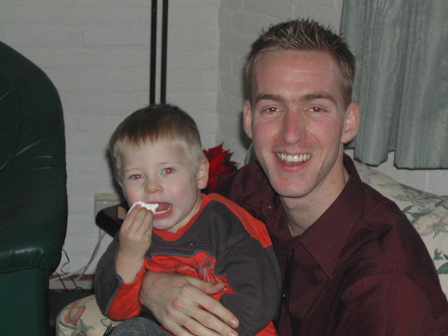 A man smiles while holding a young child who is eating a snack.