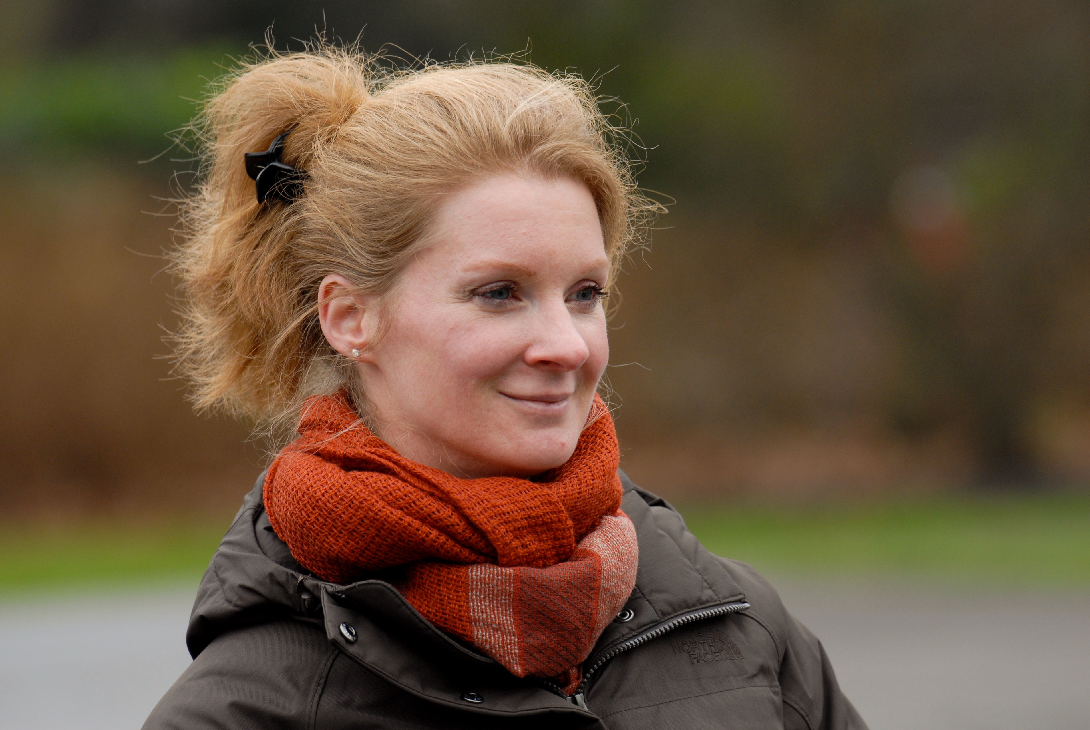 A woman with light skin and blonde hair smiles while wearing a brown jacket and an orange scarf outdoors.