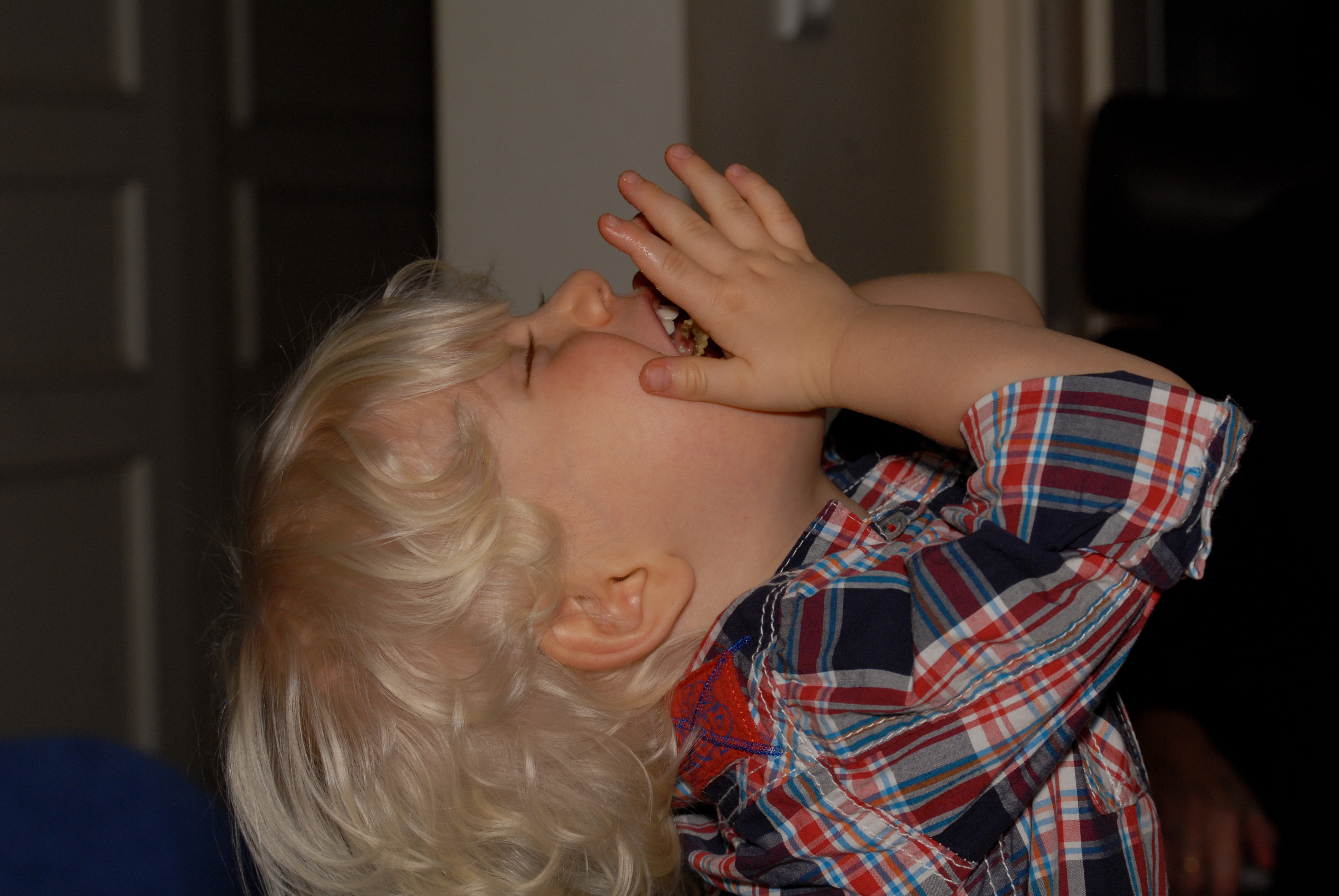 A young child with blonde hair and a plaid shirt eats a treat with both hands, head tilted back.