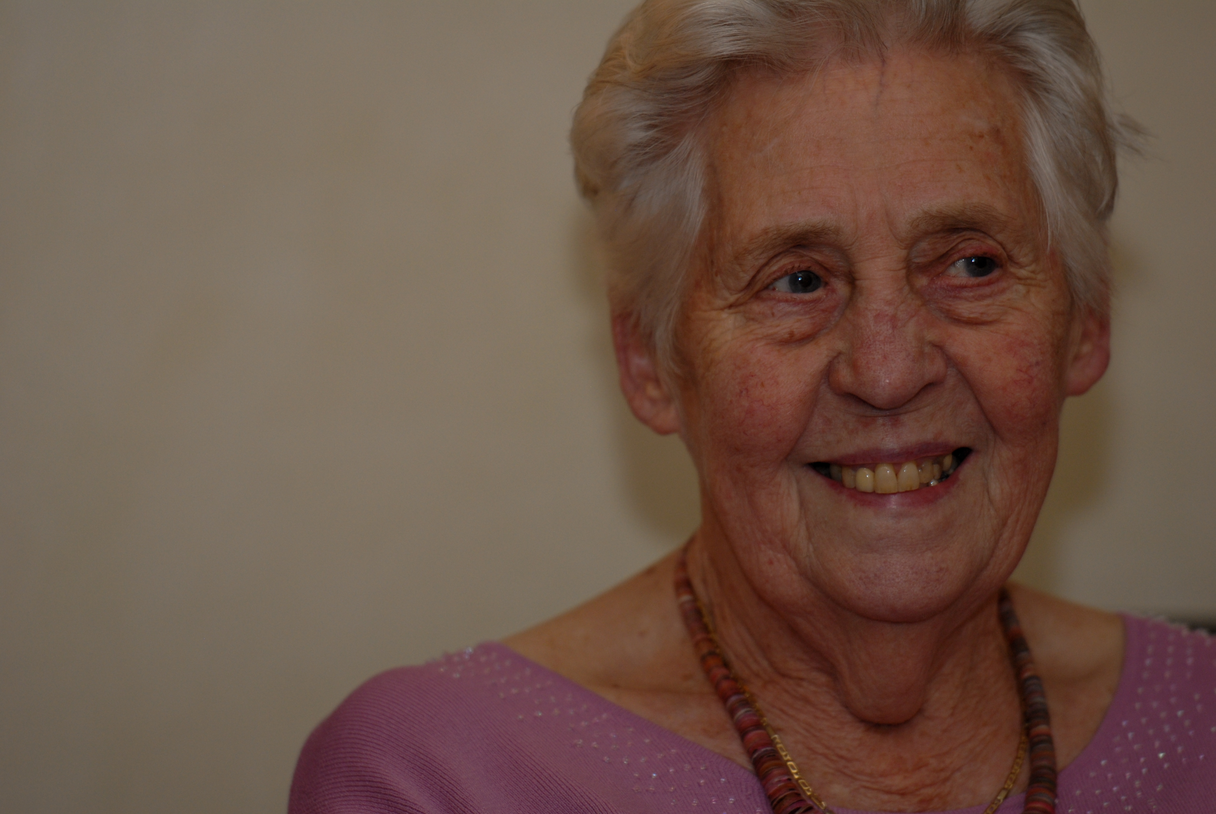An elderly woman with short white hair smiles while wearing a pink top and a beaded necklace.