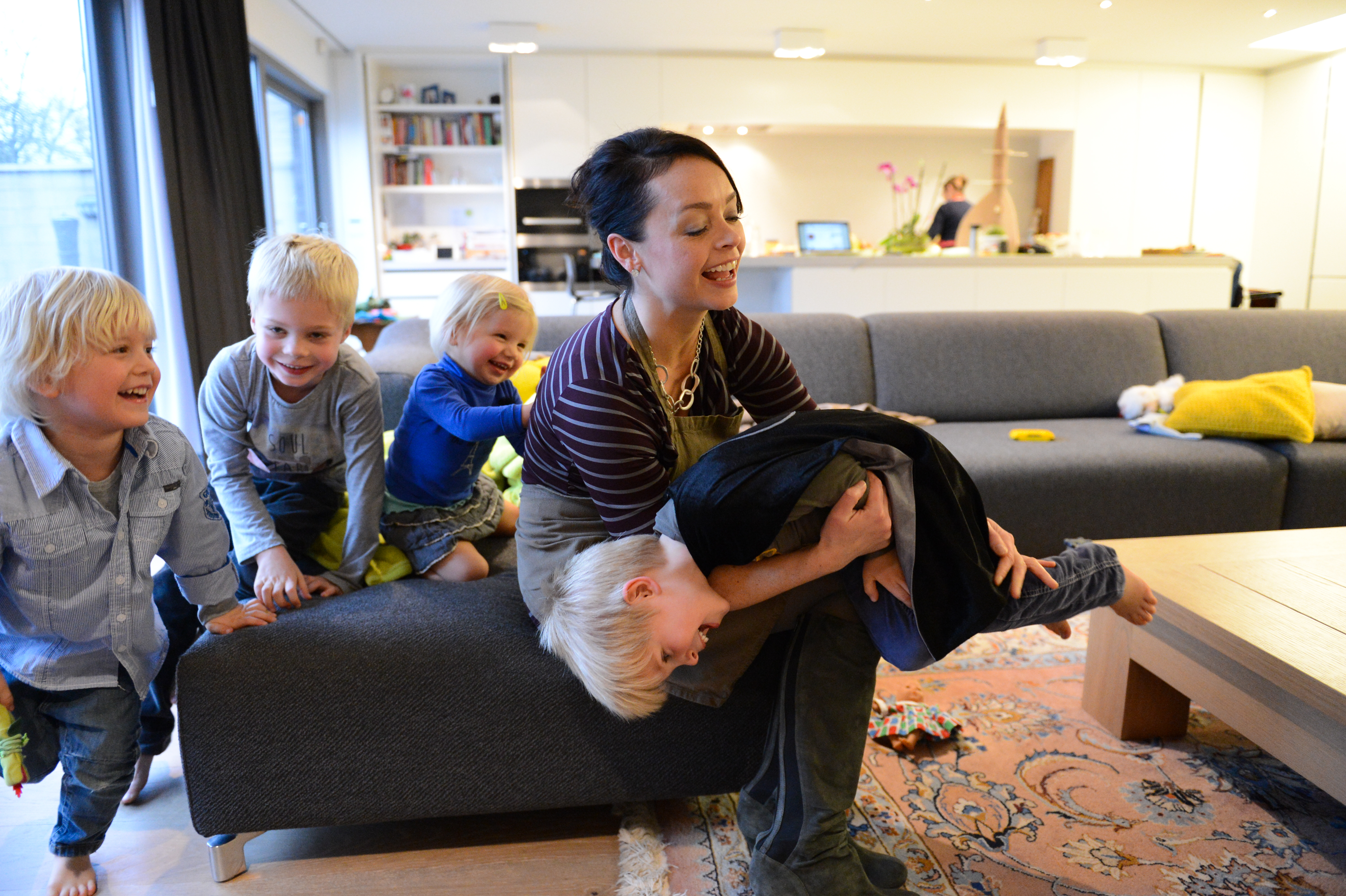 A woman playfully holds a child upside down while other children laugh and watch from the couch.