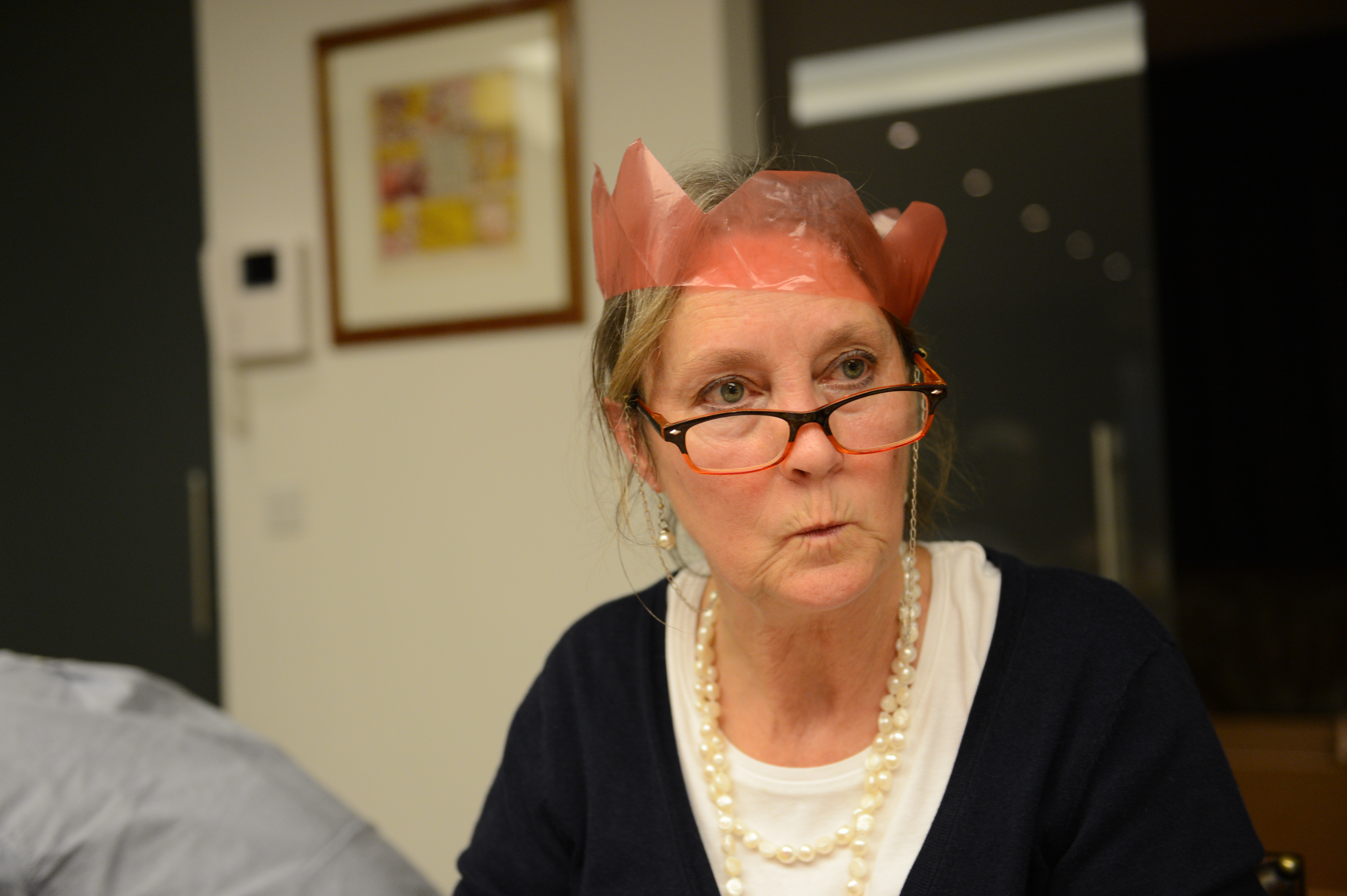 A woman wearing glasses and a red paper crown looks surprised or focused during a Christmas Eve gathering.