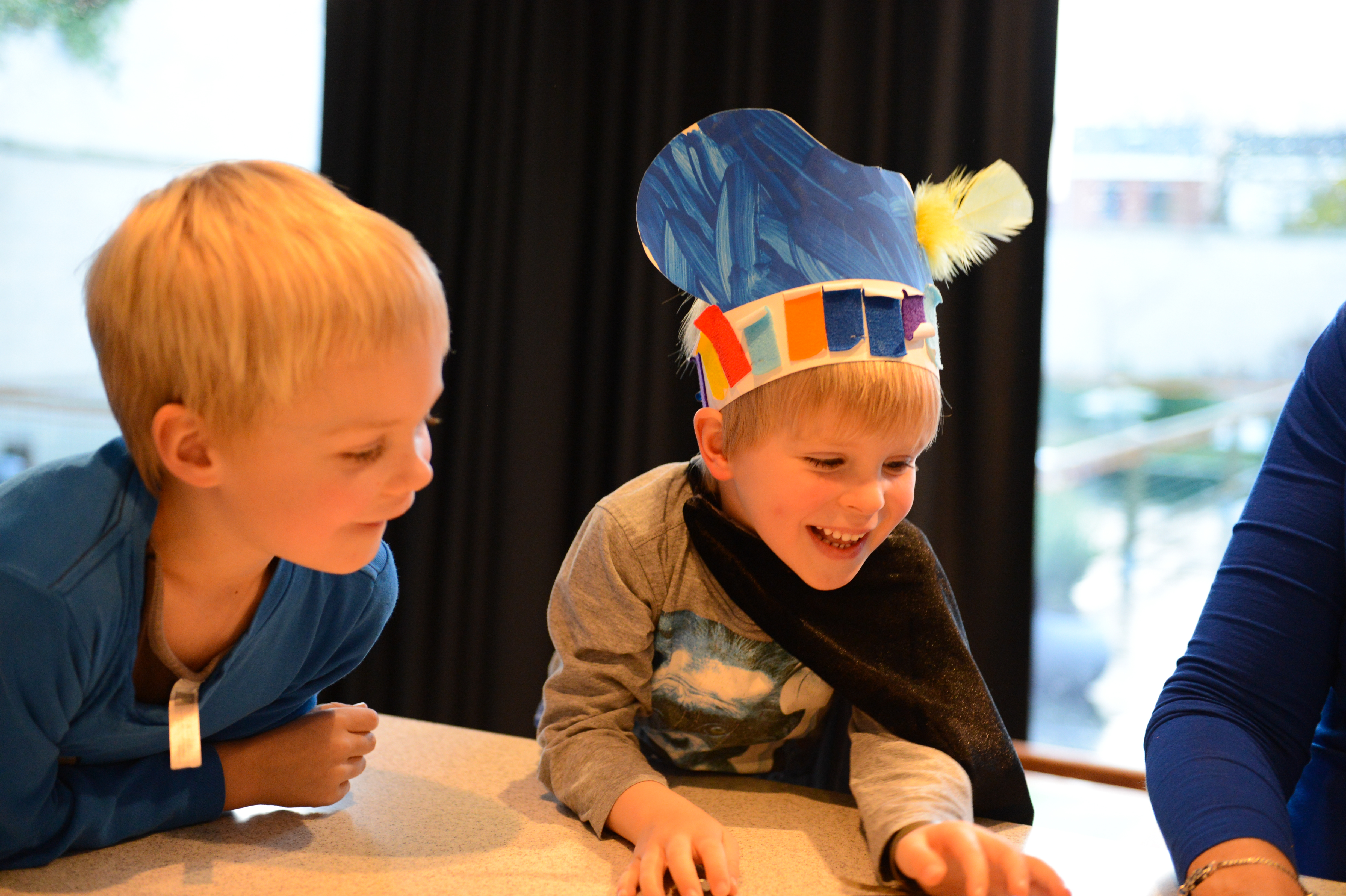 Two young children play at a table, one wearing a colorful handmade hat and smiling excitedly.