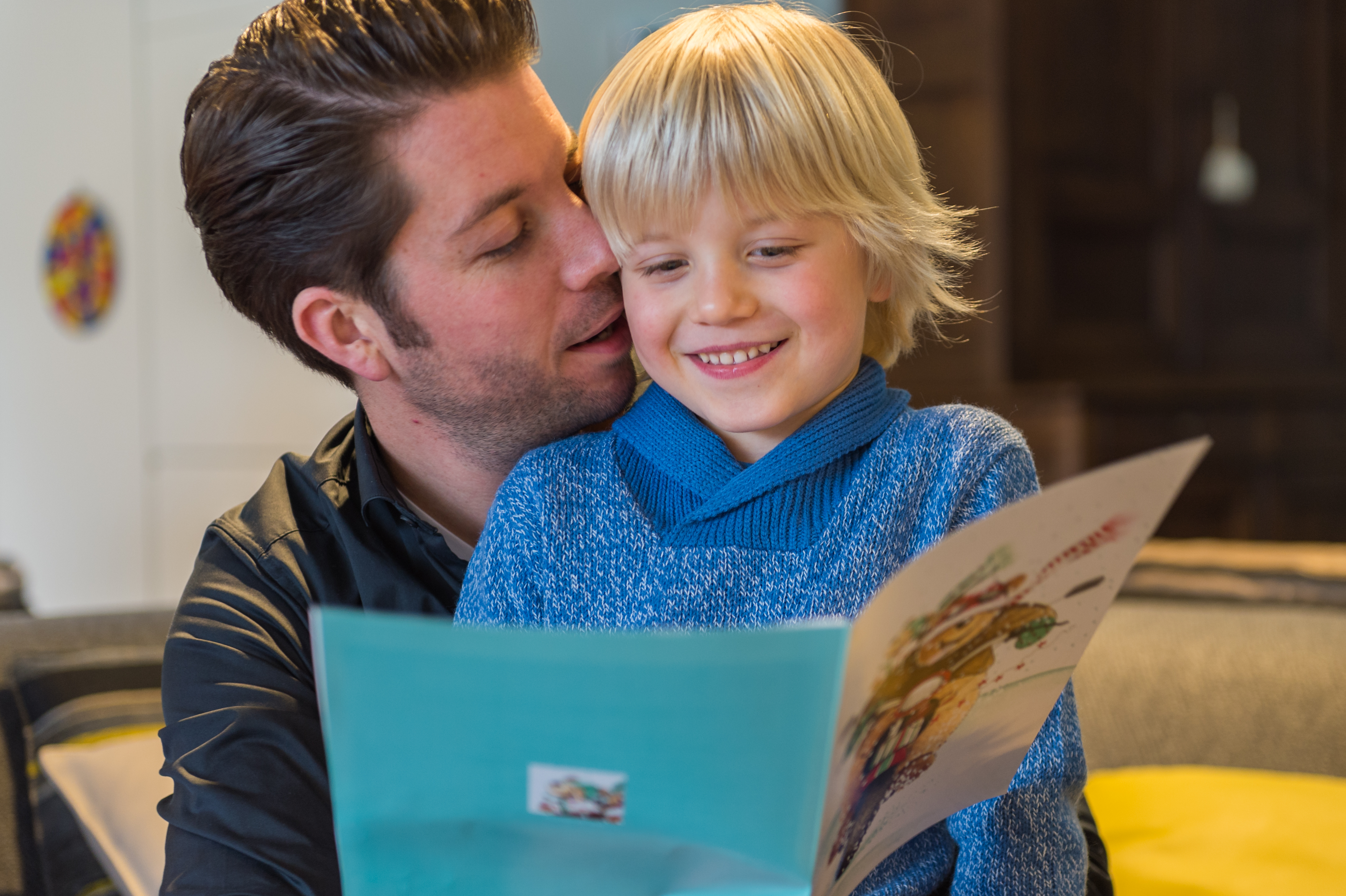 A smiling child in a blue sweater reads a Christmas card while sitting with an adult who is leaning in.