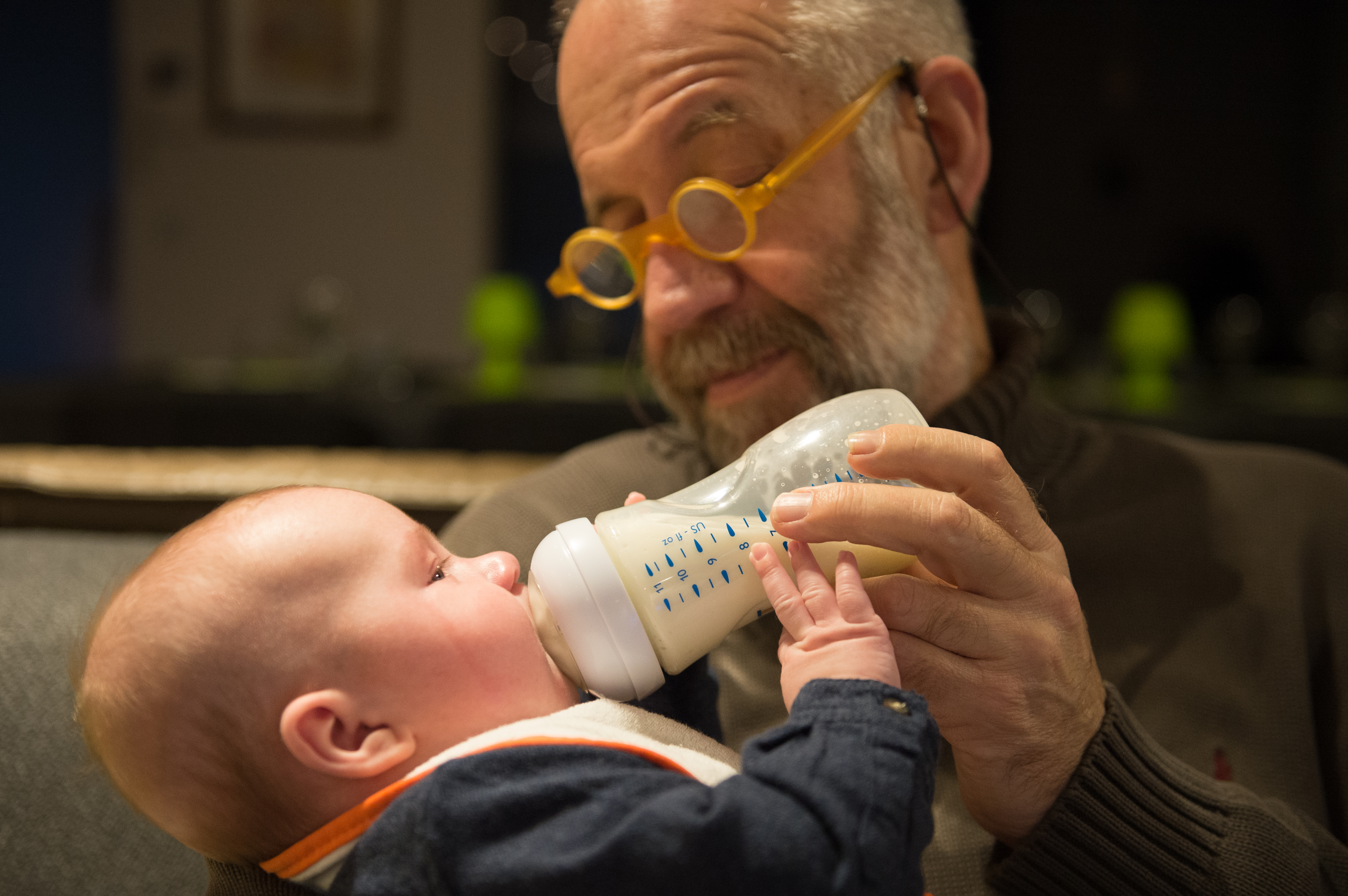 An older man with glasses feeds a baby from a bottle while sitting together on a couch.
