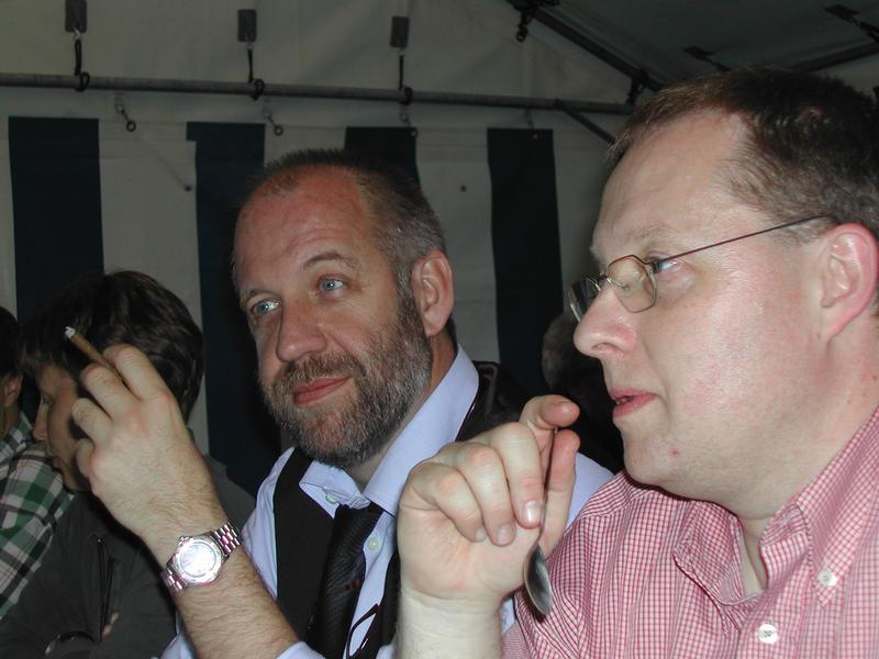 Two men sitting at a table, one holding a cigarette and the other holding a spoon, engaged in conversation.