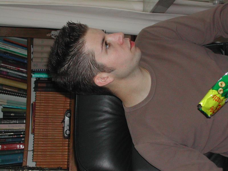 A young man in a brown shirt sits on a black chair, holding a snack bag on his lap.