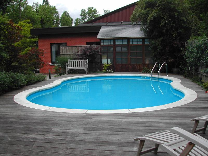 A backyard swimming pool with a wooden deck, lounge chairs, and a bench near a red brick house.