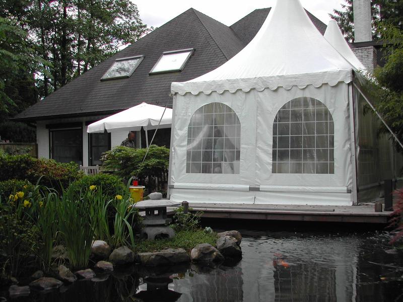 A white event tent is set up in a backyard near a pond, with a person standing nearby.
