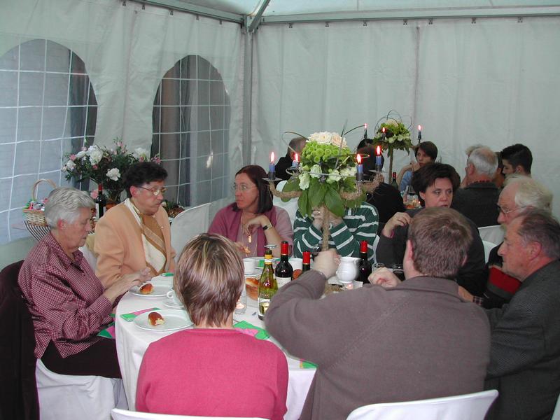 People gathered around a table in a tent, eating and talking during a communion celebration.