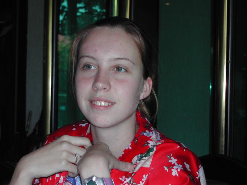 A young woman in a red floral shirt sits indoors, smiling and looking slightly away from the camera.