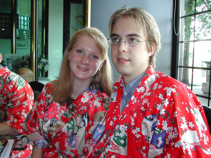 Two people wearing matching red floral-patterned robes sit together, smiling at the camera in an indoor setting.