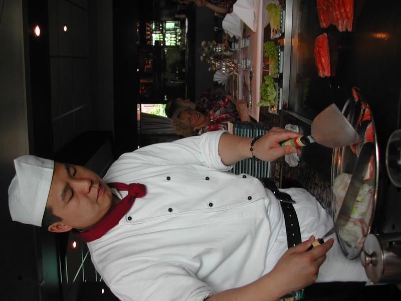 A chef in a white uniform prepares food on a hot grill while guests watch from a table.