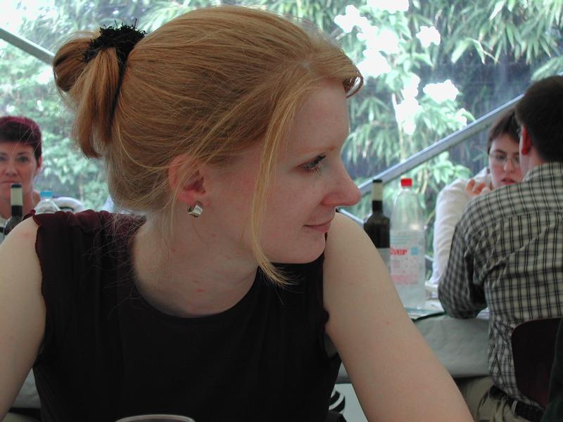 A woman with tied-back hair sits at a table, looking to the side, with people talking in the background.
