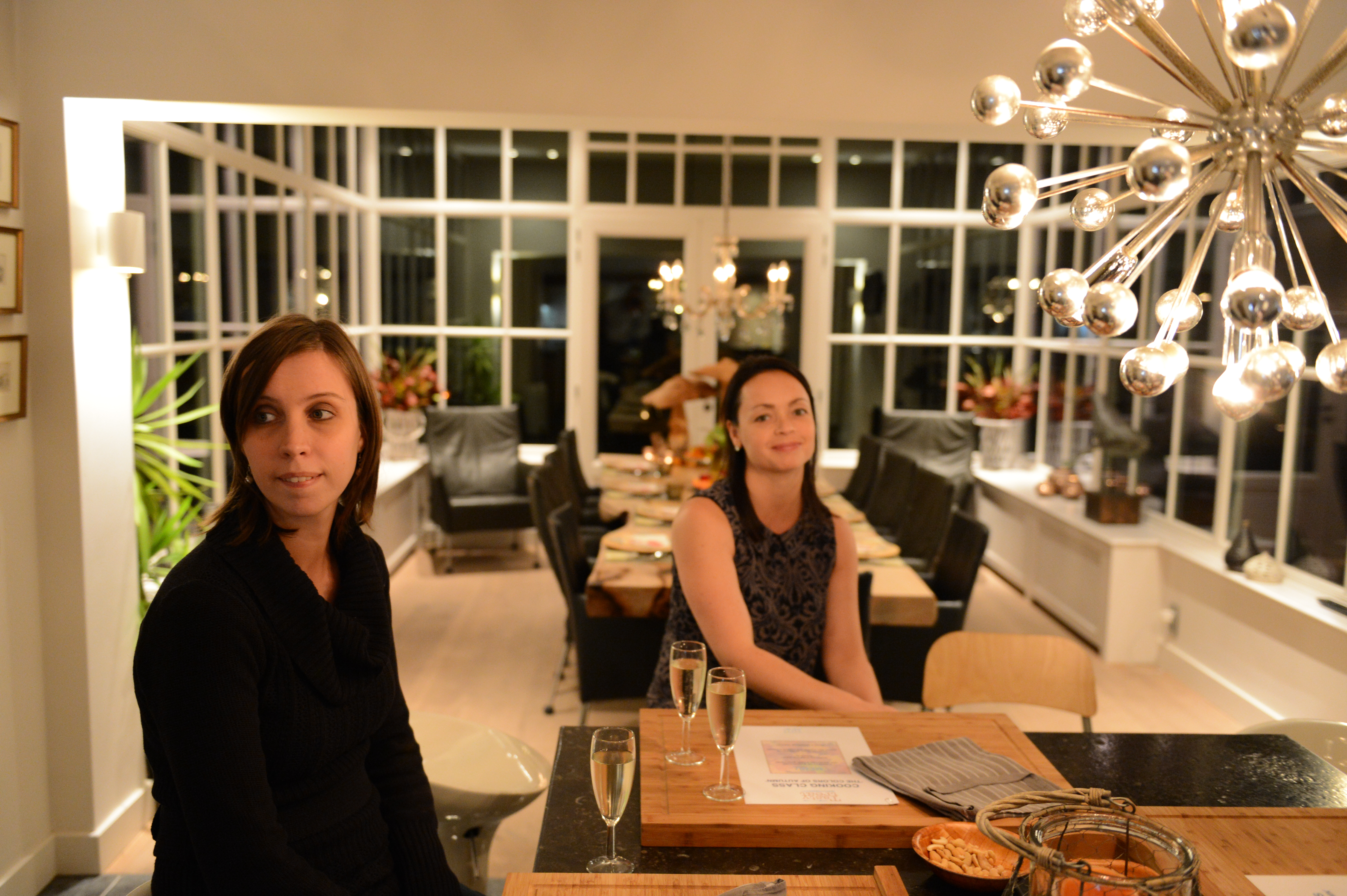 Two women sit at a kitchen counter with drinks, waiting for a cooking class to begin.