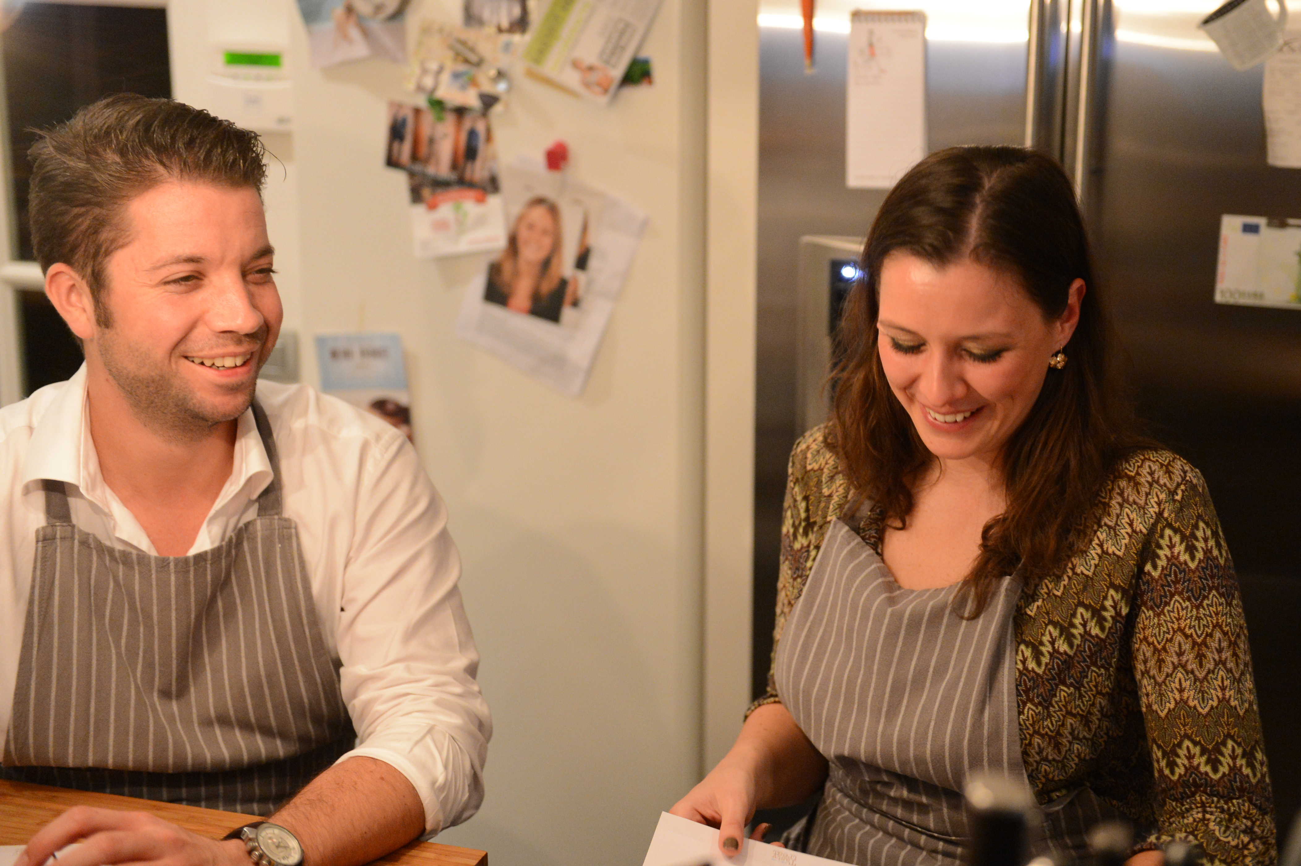 Two people wearing aprons smile while participating in a cooking class, looking at a piece of paper.