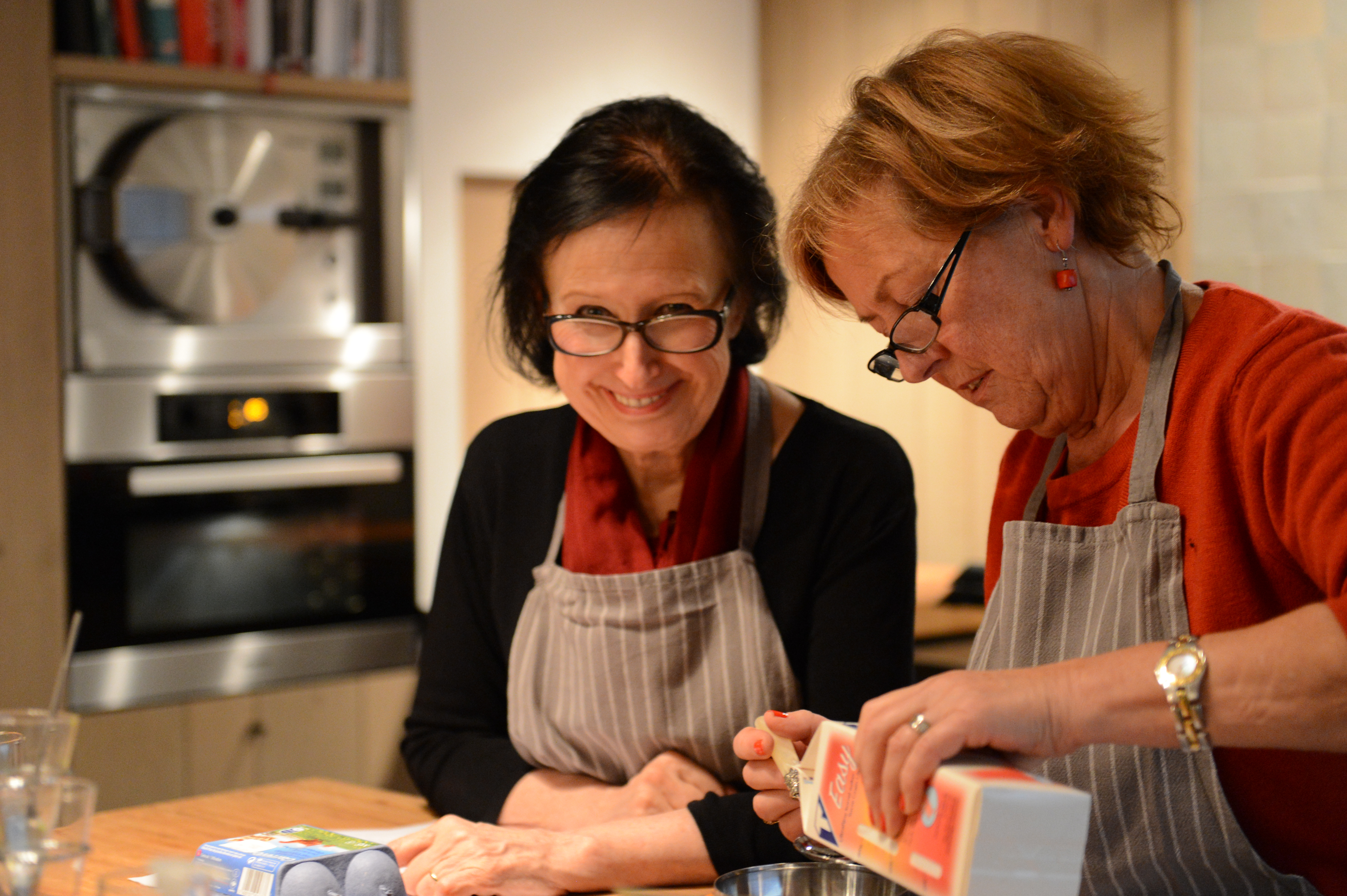 Two women in aprons participate in a cooking class, one pouring an ingredient into a bowl while the other smiles.