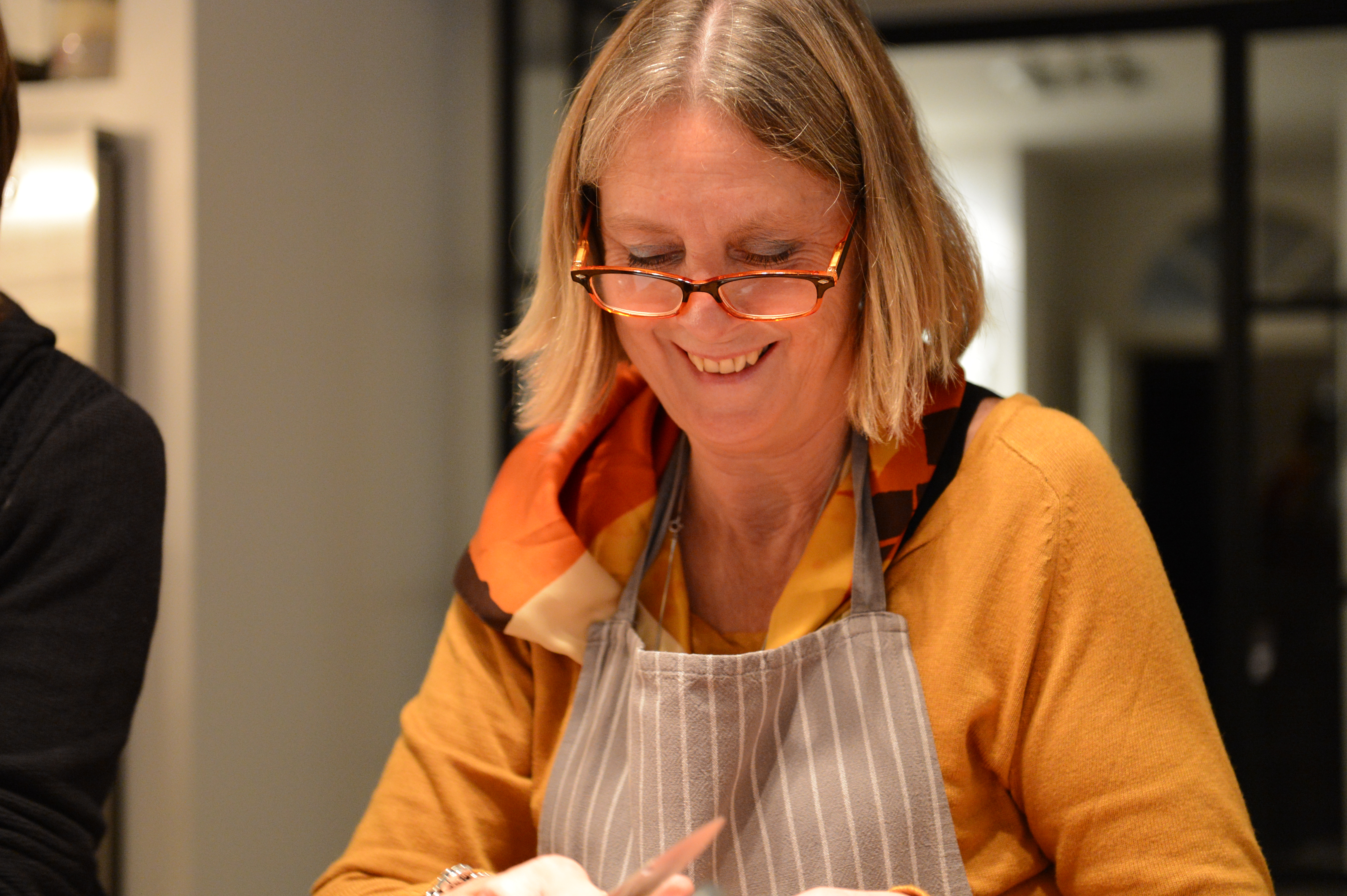 A woman wearing an apron and glasses smiles while preparing food during a cooking class.