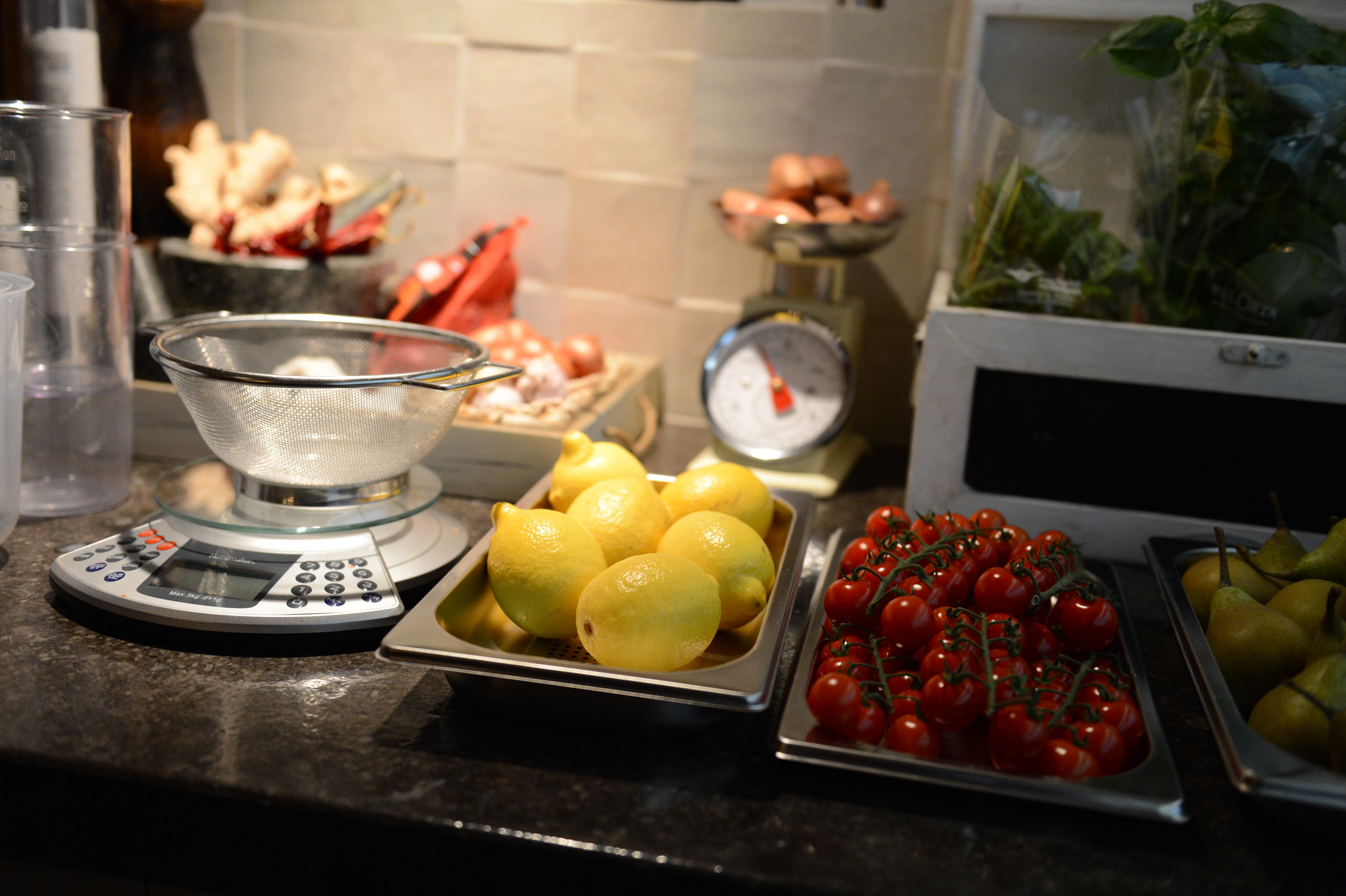 Fresh lemons, cherry tomatoes, and pears on trays beside a kitchen scale and strainer during a cooking class.