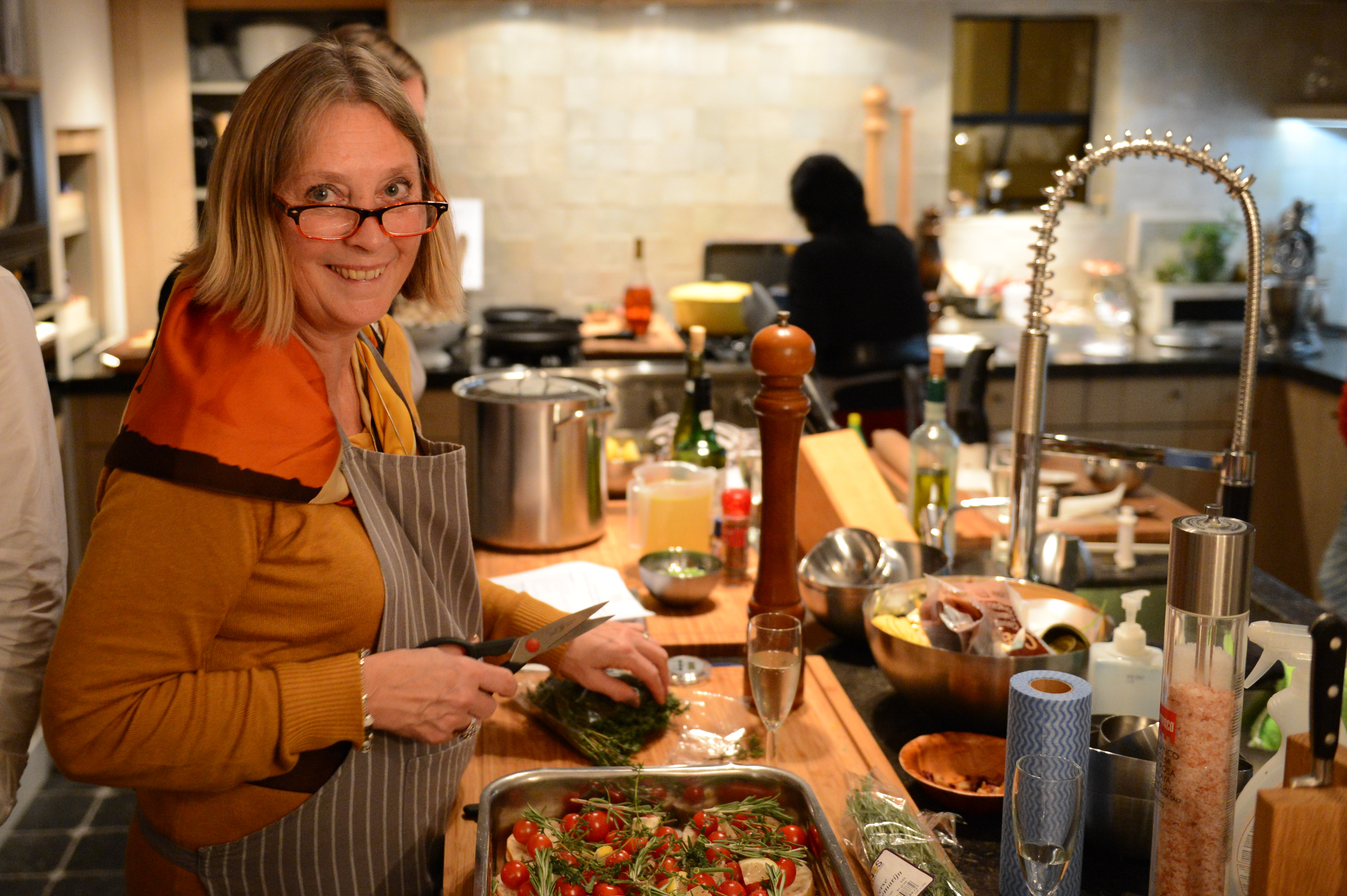 A woman in an apron smiles while chopping herbs in a busy kitchen during a cooking class.