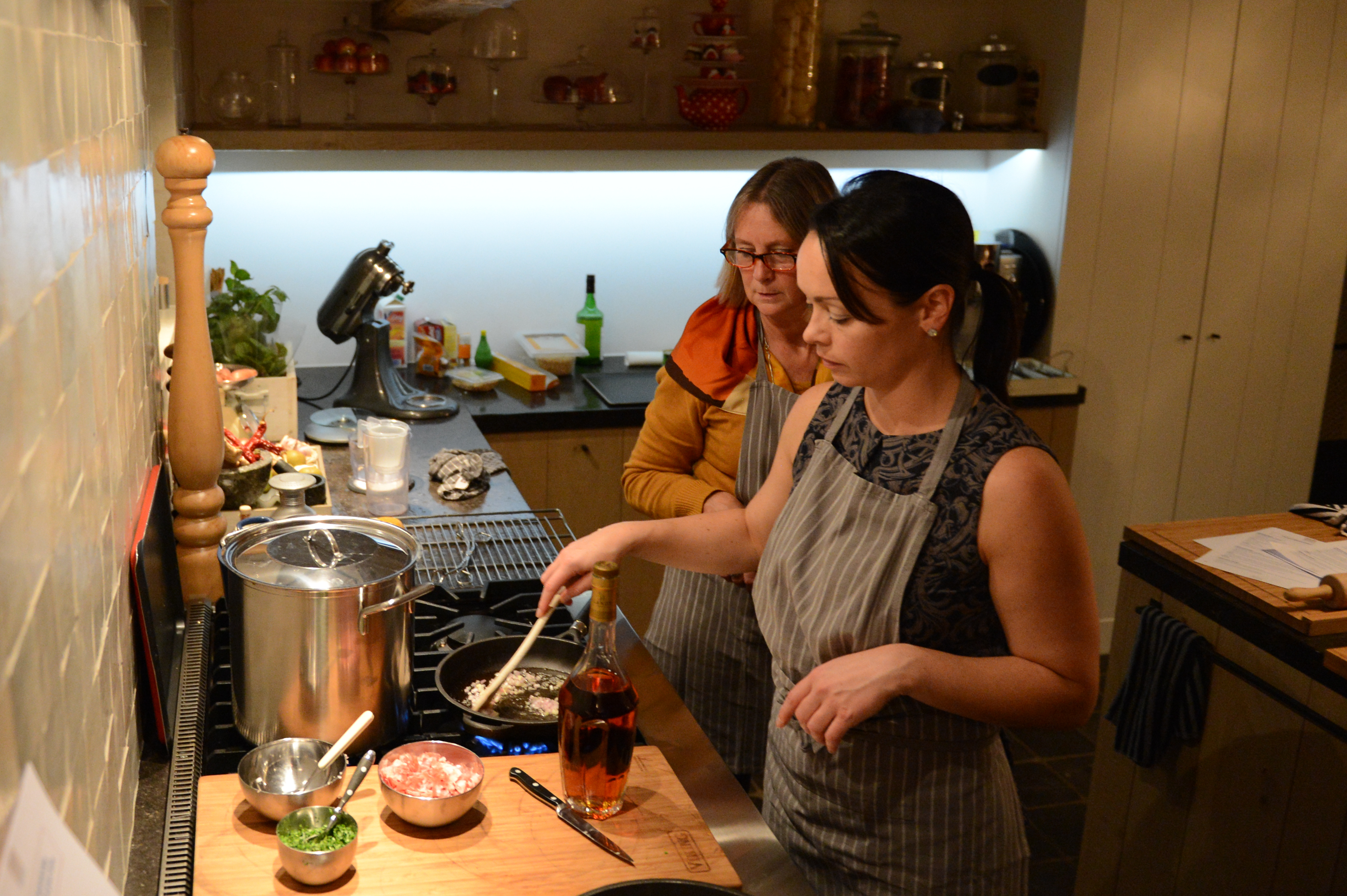 Two women in aprons cooking at a stove, one stirring a pan while the other observes.