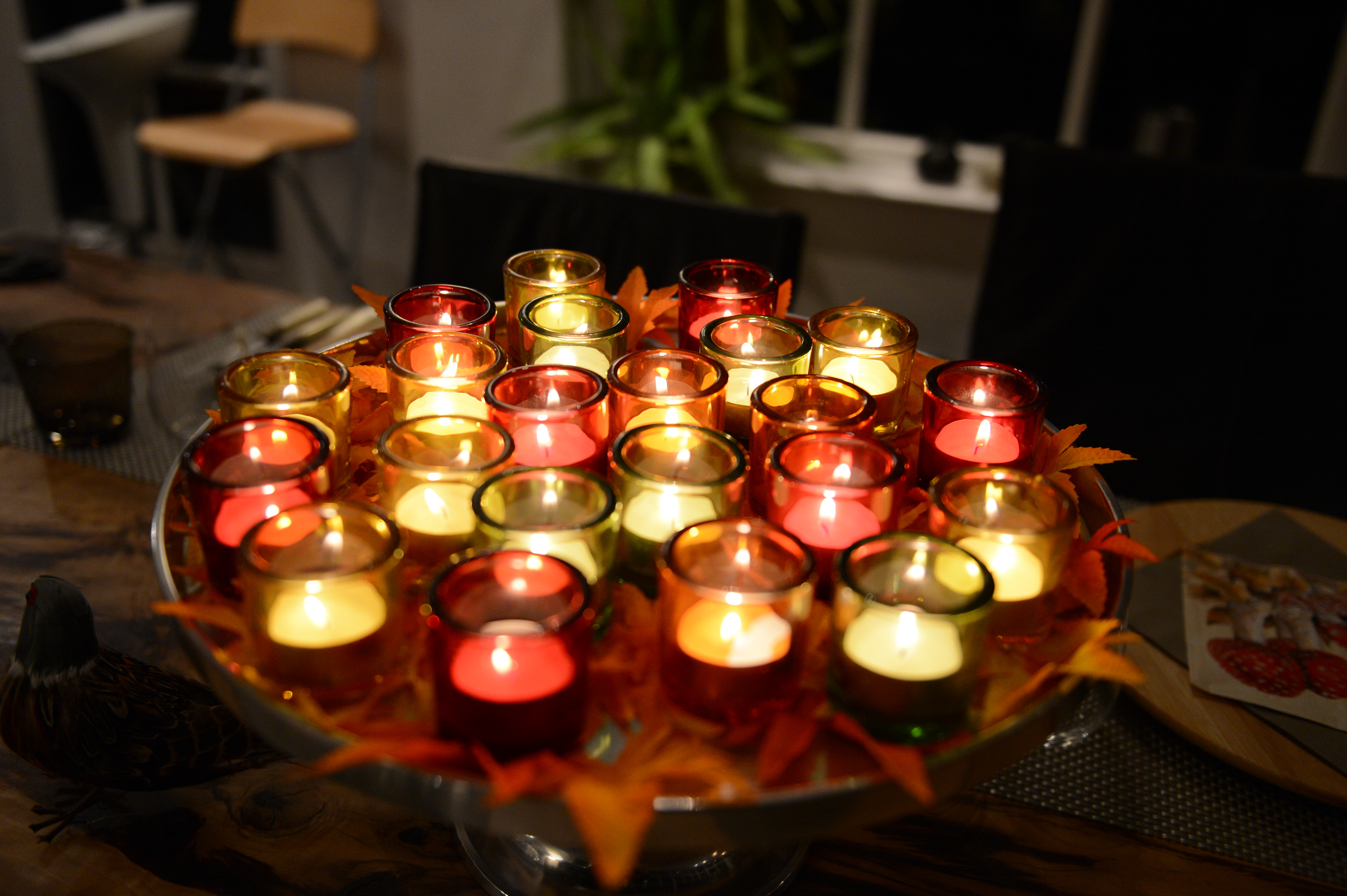 A table centerpiece with multiple lit candles in red and yellow glass holders, decorated with autumn leaves.