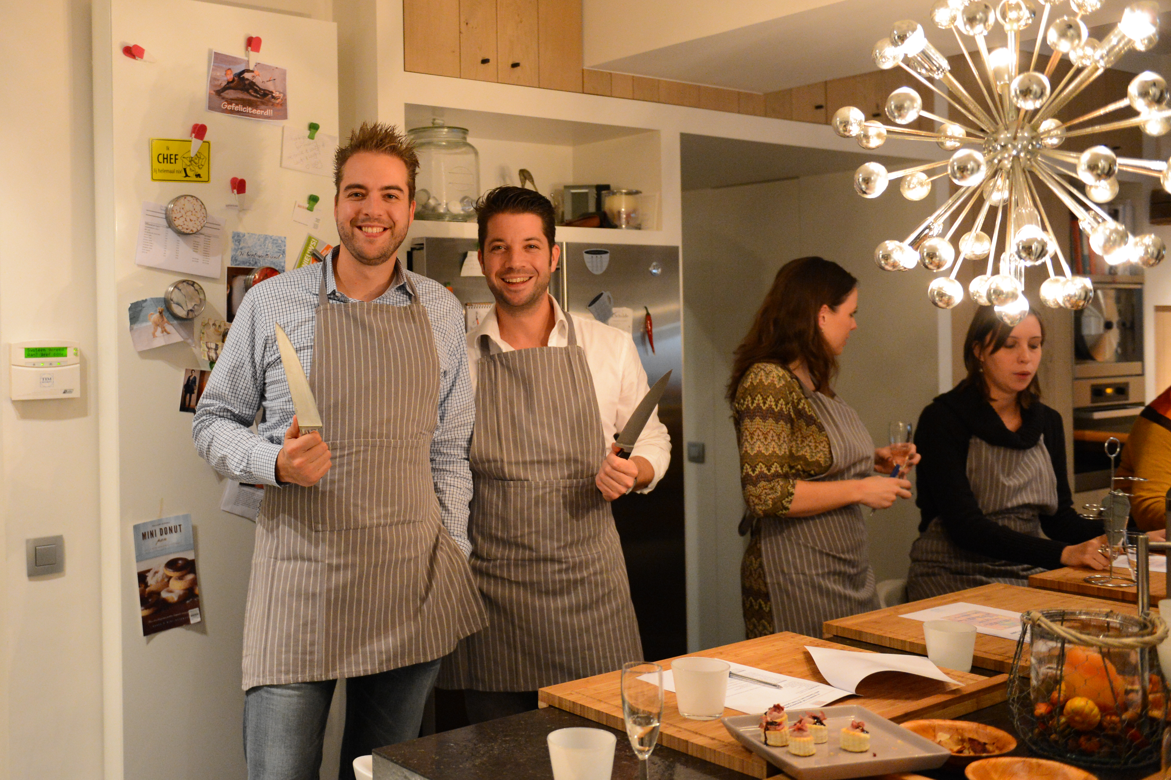 Two smiling men in aprons hold knives, posing for the camera during a cooking class.