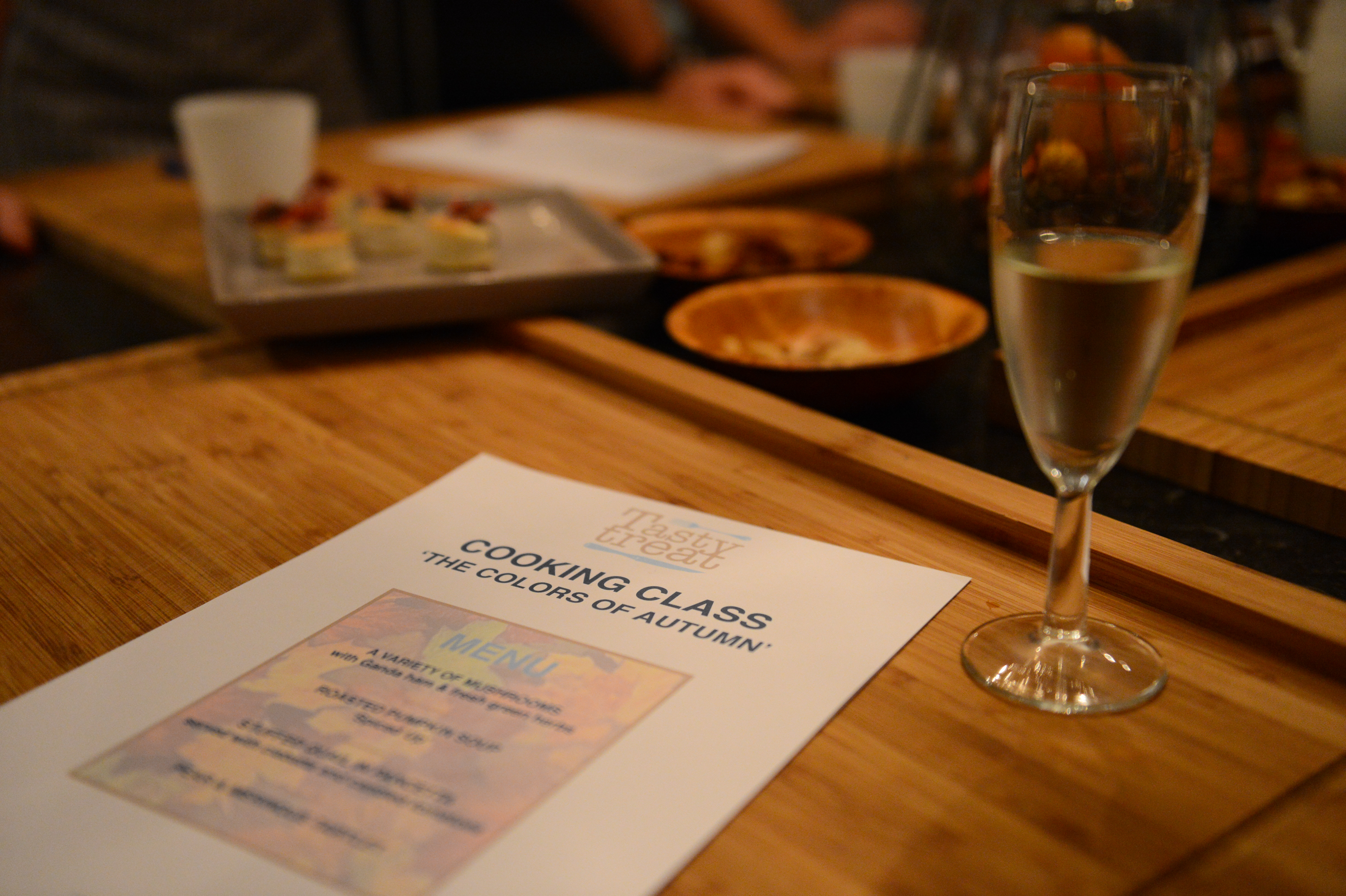 A cooking class menu on a wooden table with a glass of champagne and plated appetizers in the background.