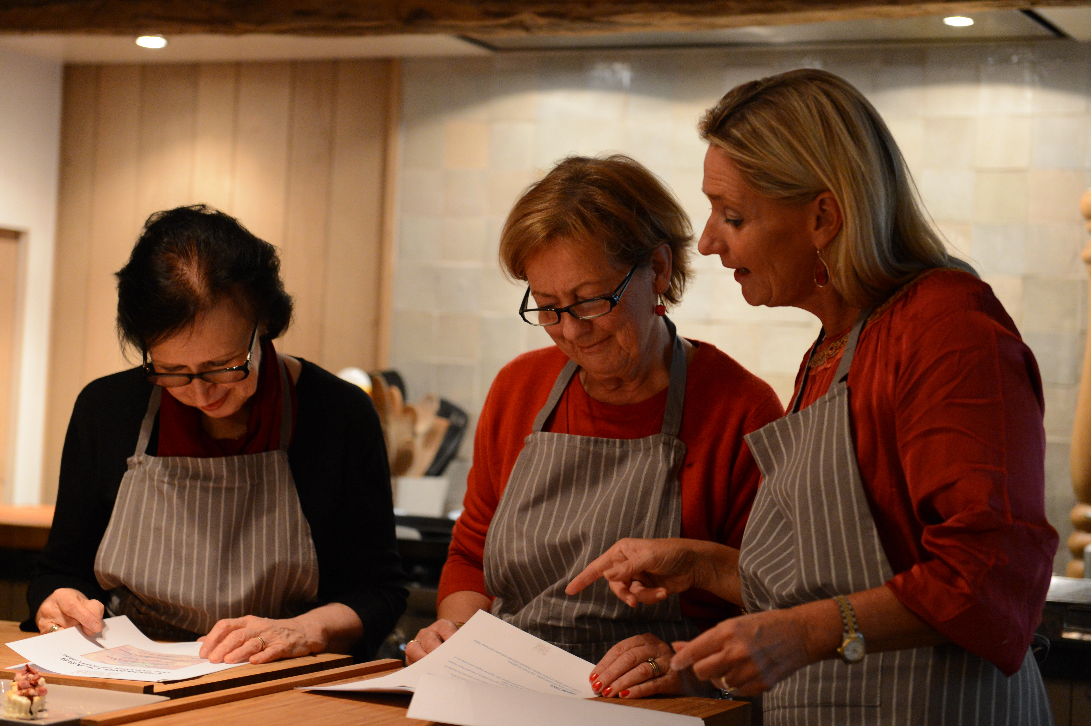 Three women in aprons review a recipe together during a cooking class, with one pointing at the paper.