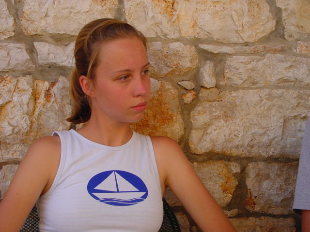 A young woman in a white tank top with a sailboat design sits against a stone wall, looking to the side.