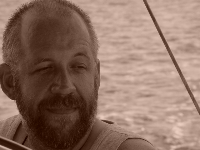 A bearded man in a tank top smiles while looking to the side, with water in the background.