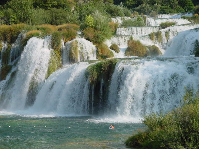Large waterfalls cascade over rocks into a river, with a person wearing a red life vest swimming below.