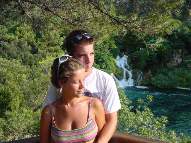 A young couple stands by a wooden railing, looking into the distance, with a waterfall and greenery behind them.