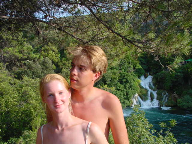 A young couple poses outdoors with a waterfall and lush greenery in the background.