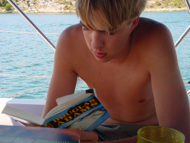 A shirtless young man sits on a boat, reading a Douglas Adams book near the water.