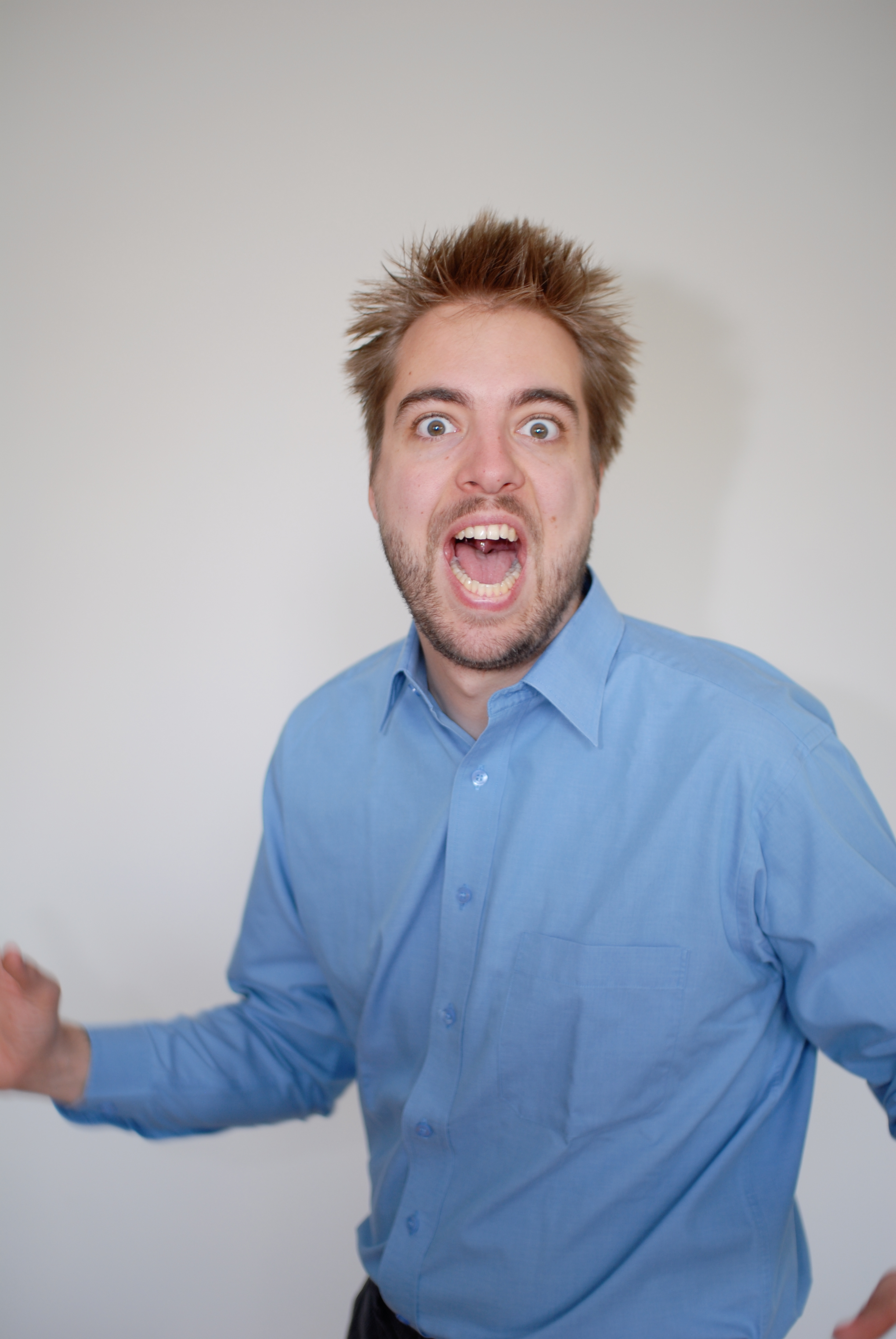 A man in a blue shirt with messy hair shouts with his mouth open and arms slightly raised.