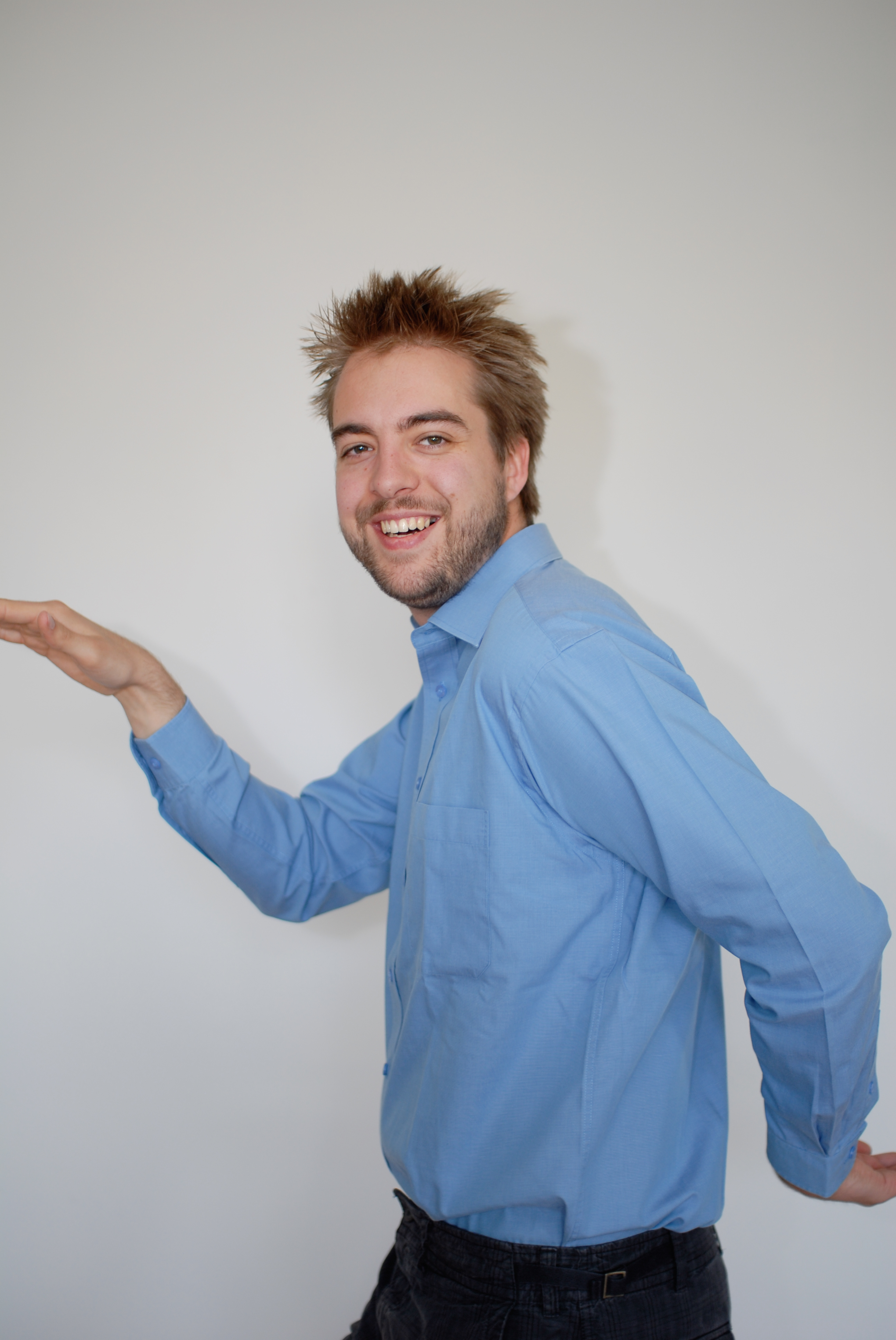 A man in a blue shirt smiles while playfully posing with his arms bent and hands raised.