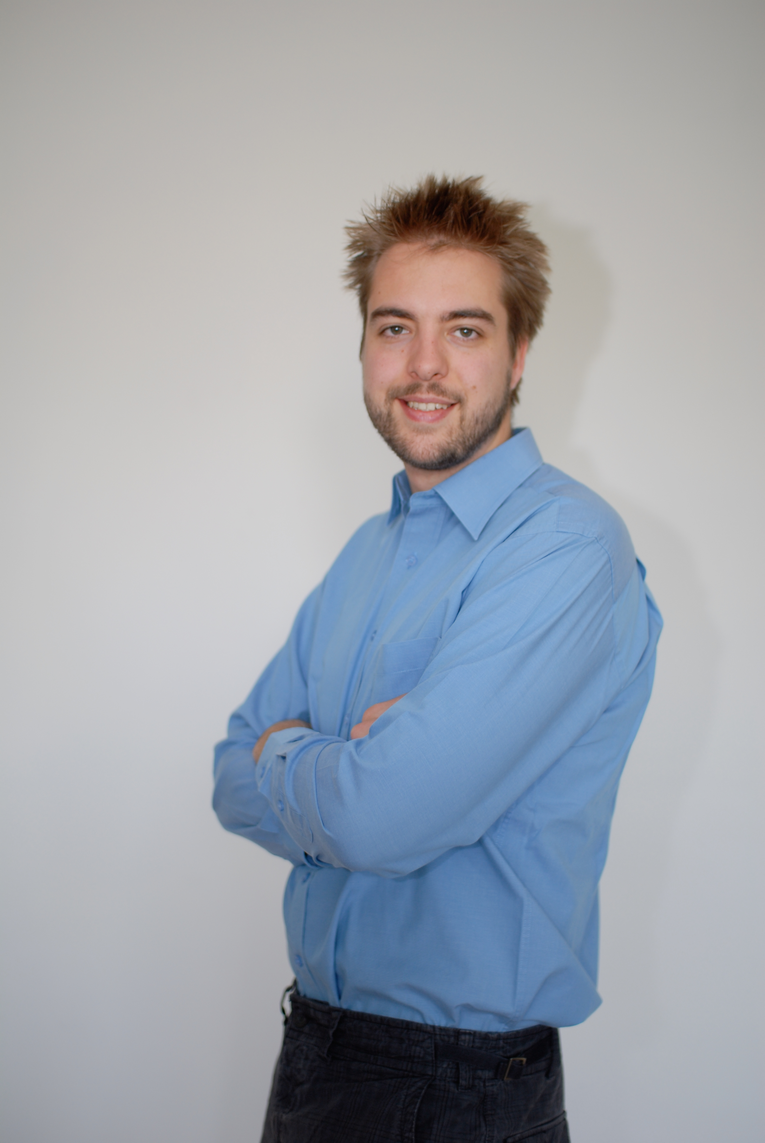 A man in a blue shirt stands against a plain background, smiling with his arms crossed.