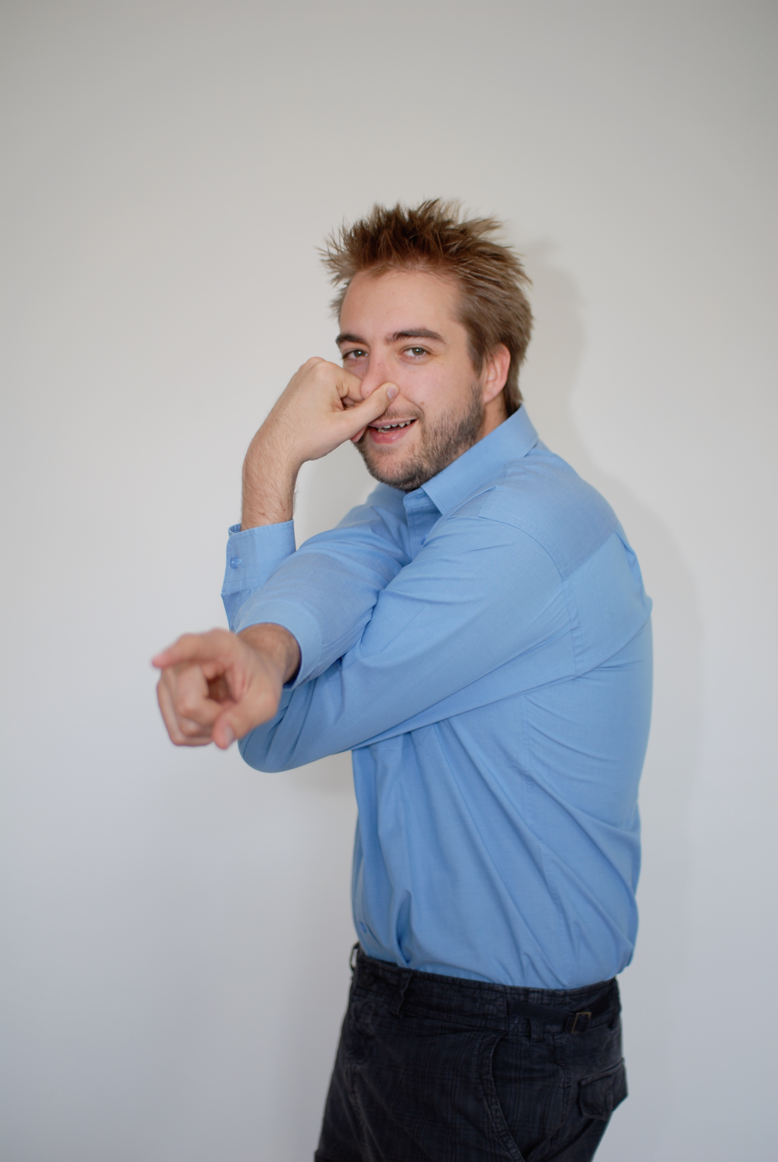 A man in a blue shirt playfully poses, pointing forward and holding his nose with his other hand.