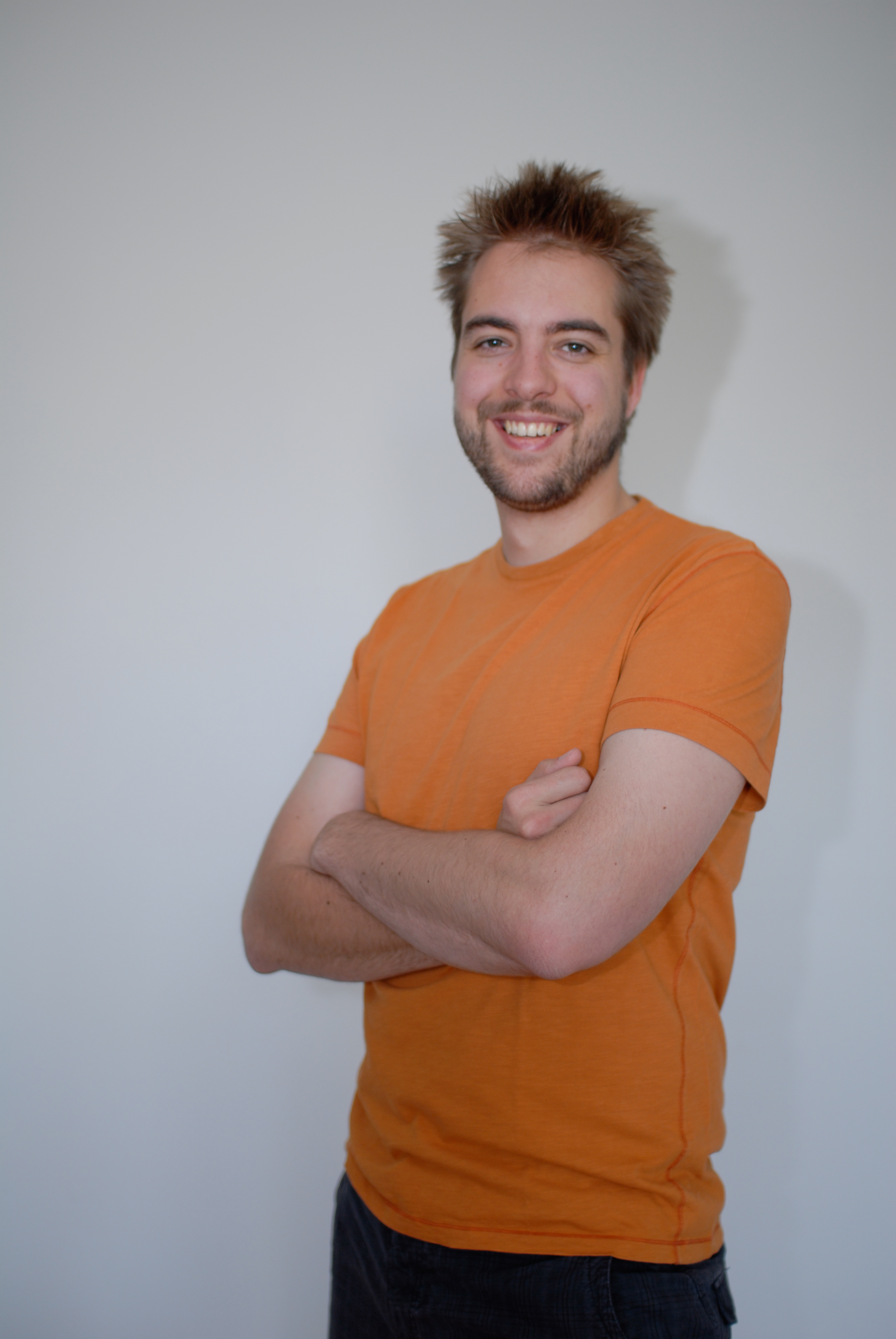 A person wearing an orange shirt stands against a plain background, smiling with arms crossed.