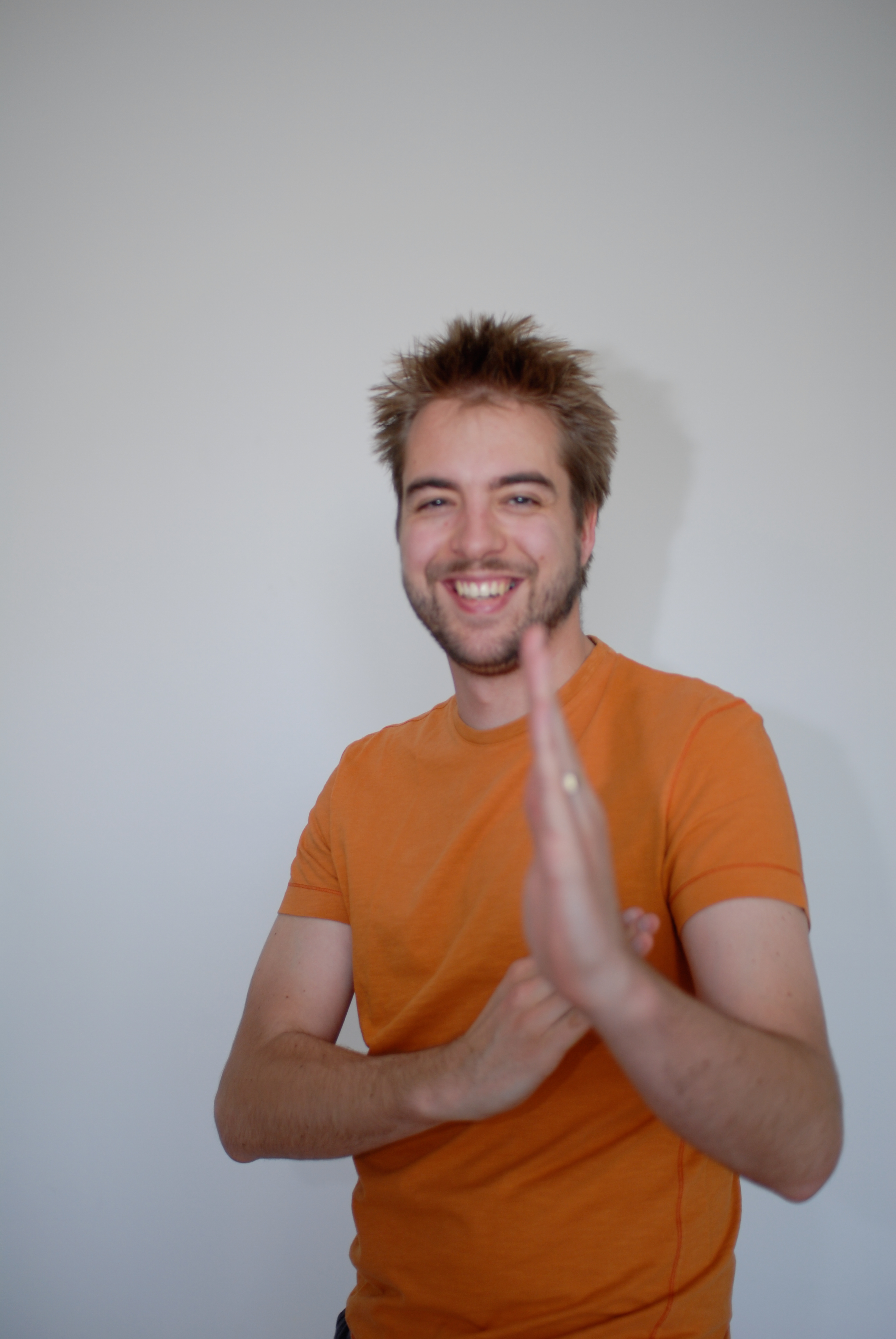 A smiling man in an orange shirt poses playfully with one hand raised in front of the camera.