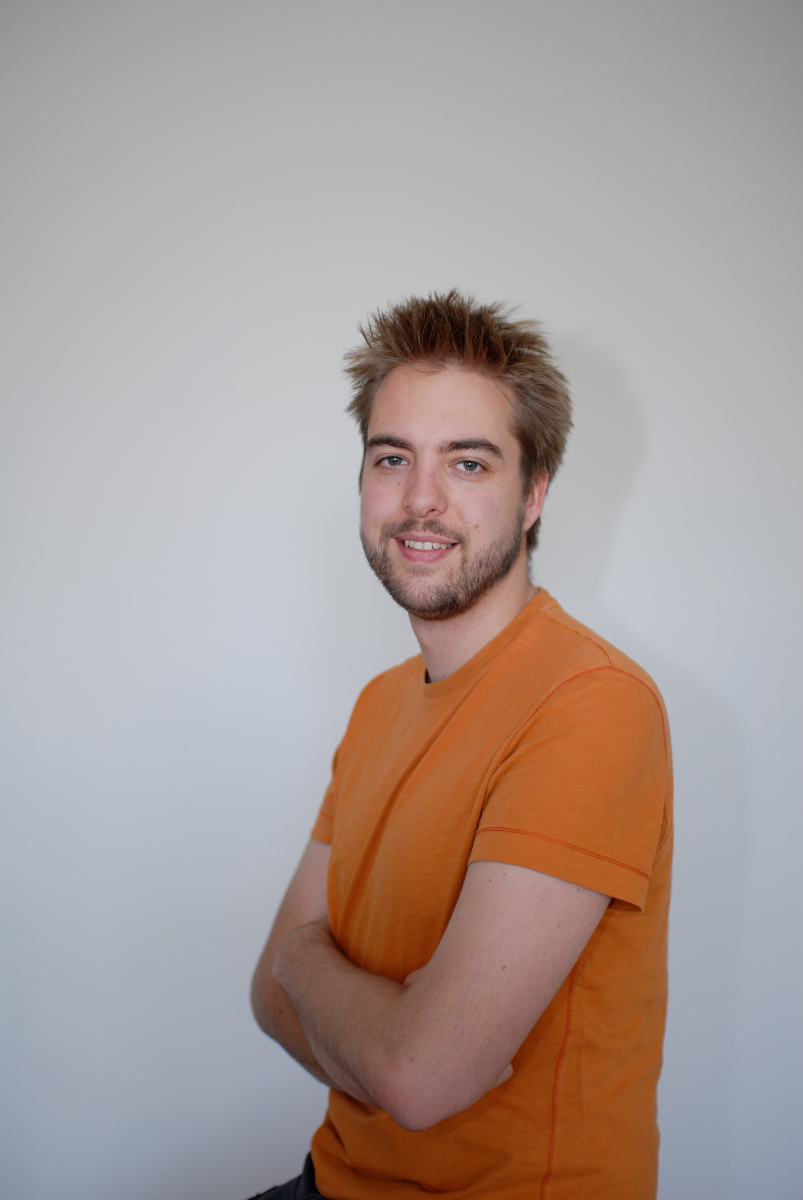 A man with short, spiky hair and a beard wears an orange shirt and smiles with arms crossed.